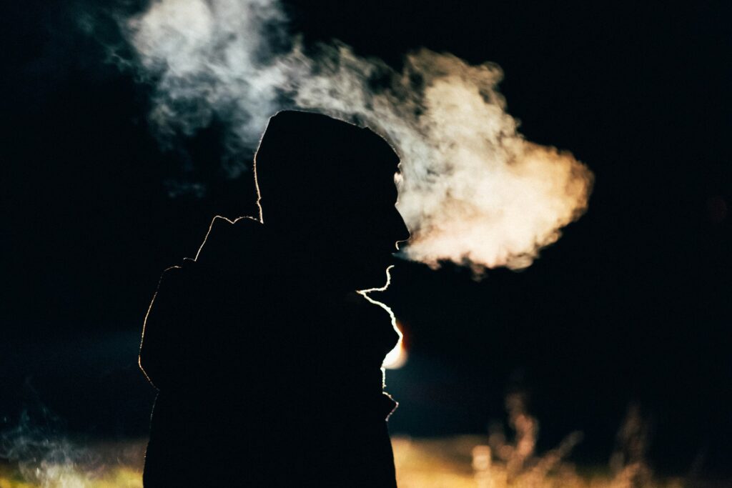 Silhouette of a person wearing a beanie on a cold night, with visible breath creating a misty cloud in the cold air against a dark background.