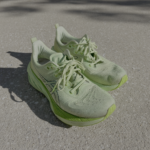 A pair of well-worn ASICS NovaBlast running shoes in bright green, placed on a concrete sidewalk. The shoes show visible signs of use, with dirt and scuff marks on the mesh upper and outsole. Shadows from the shoes are cast onto the pavement under bright sunlight.