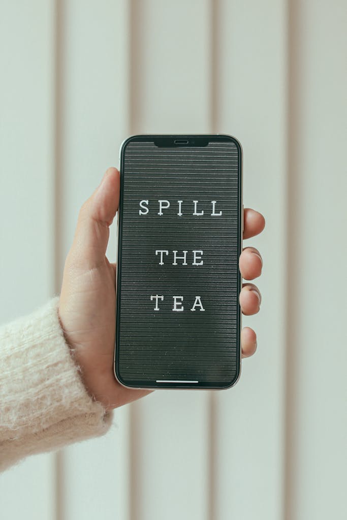 A hand holding a smartphone displaying the text 'Spill the Tea', symbolizing gossip or sharing news.